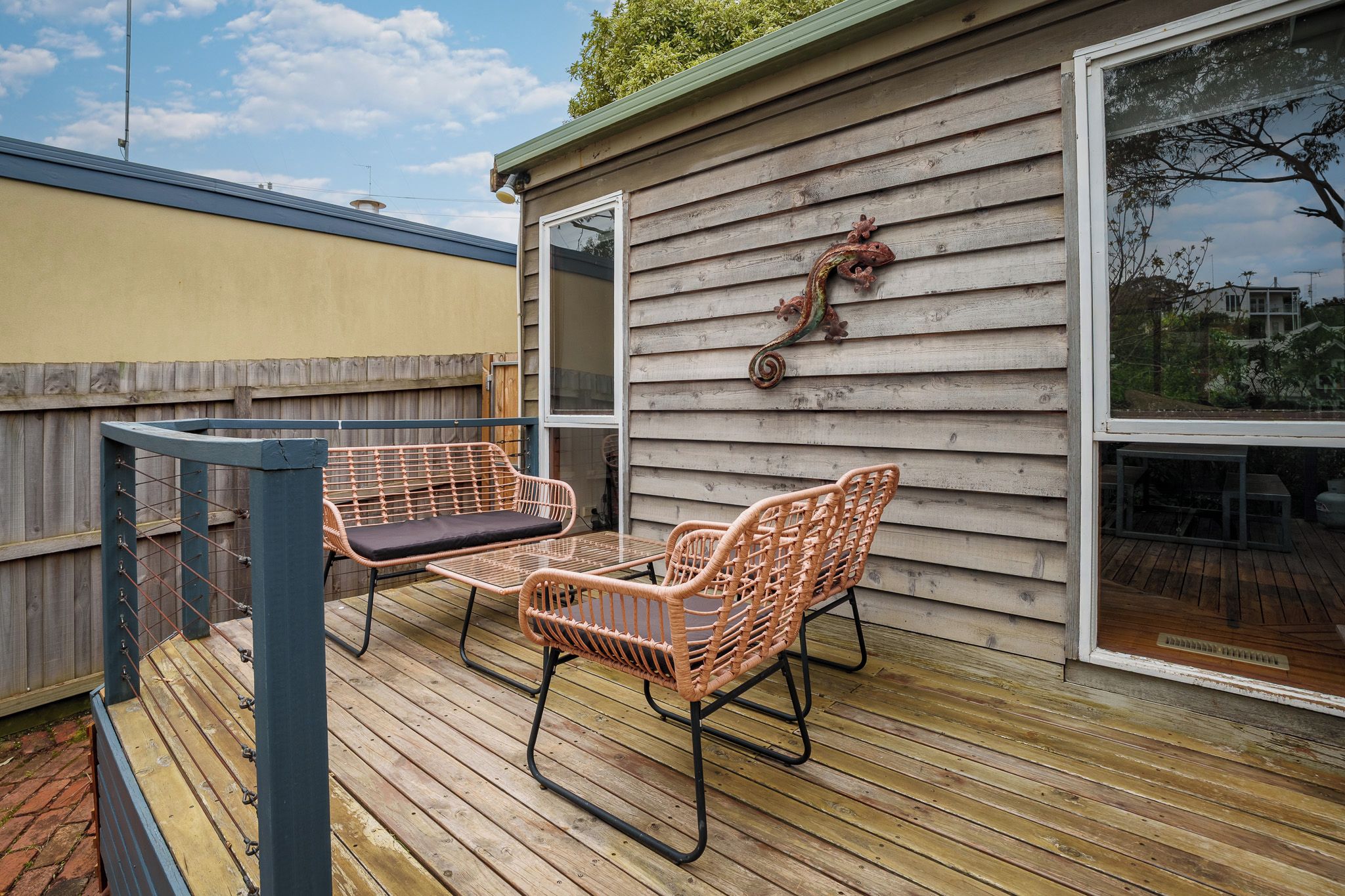 Eine Holzterrasse mit zwei Korbstühlen und einem Tisch, ein Holenzaun und ein Haus mit Fenster und einem dekorativen Element an der Wand.