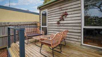Eine Holzterrasse mit zwei Korbstühlen und einem Tisch, ein Holenzaun und ein Haus mit Fenster und einem dekorativen Element an der Wand.