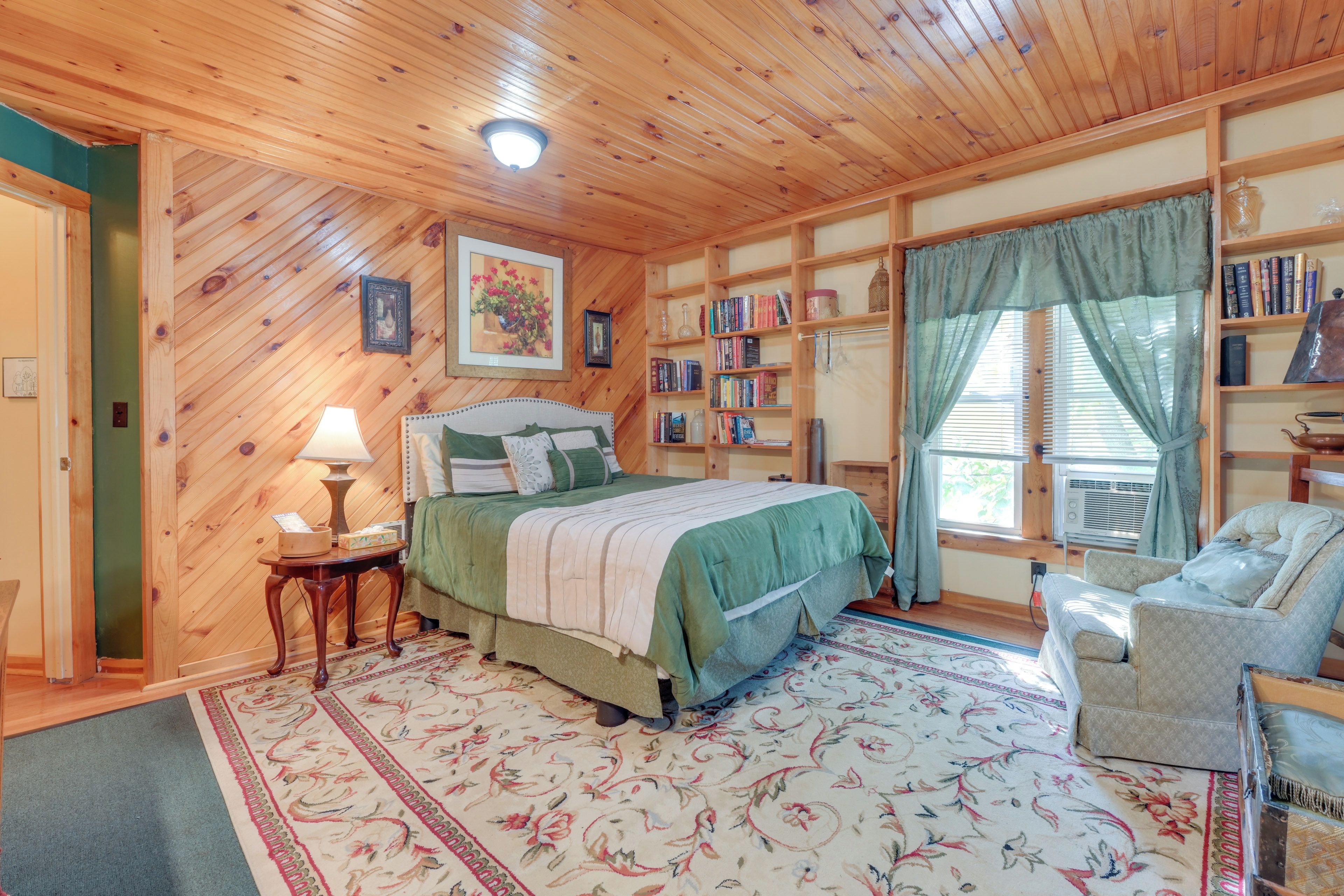 Un dormitorio acogedor con techo de madera, una cama con colcha verde, una mesita de noche con lámpara, una estantería y una ventana con cortinas.