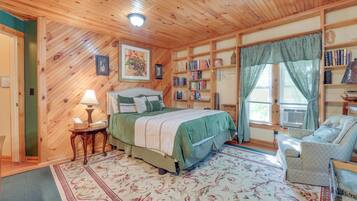 Un dormitorio acogedor con techo de madera, una cama con colcha verde, una mesita de noche con lámpara, una estantería y una ventana con cortinas.