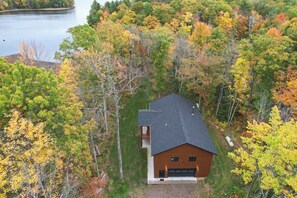 A single-story house with a dark roof, surrounded by a dense forest with autumn foliage, and a body of water in the background. - Cozy Lakefront Retreat w/ Patio on the Big Chip (Hayward)