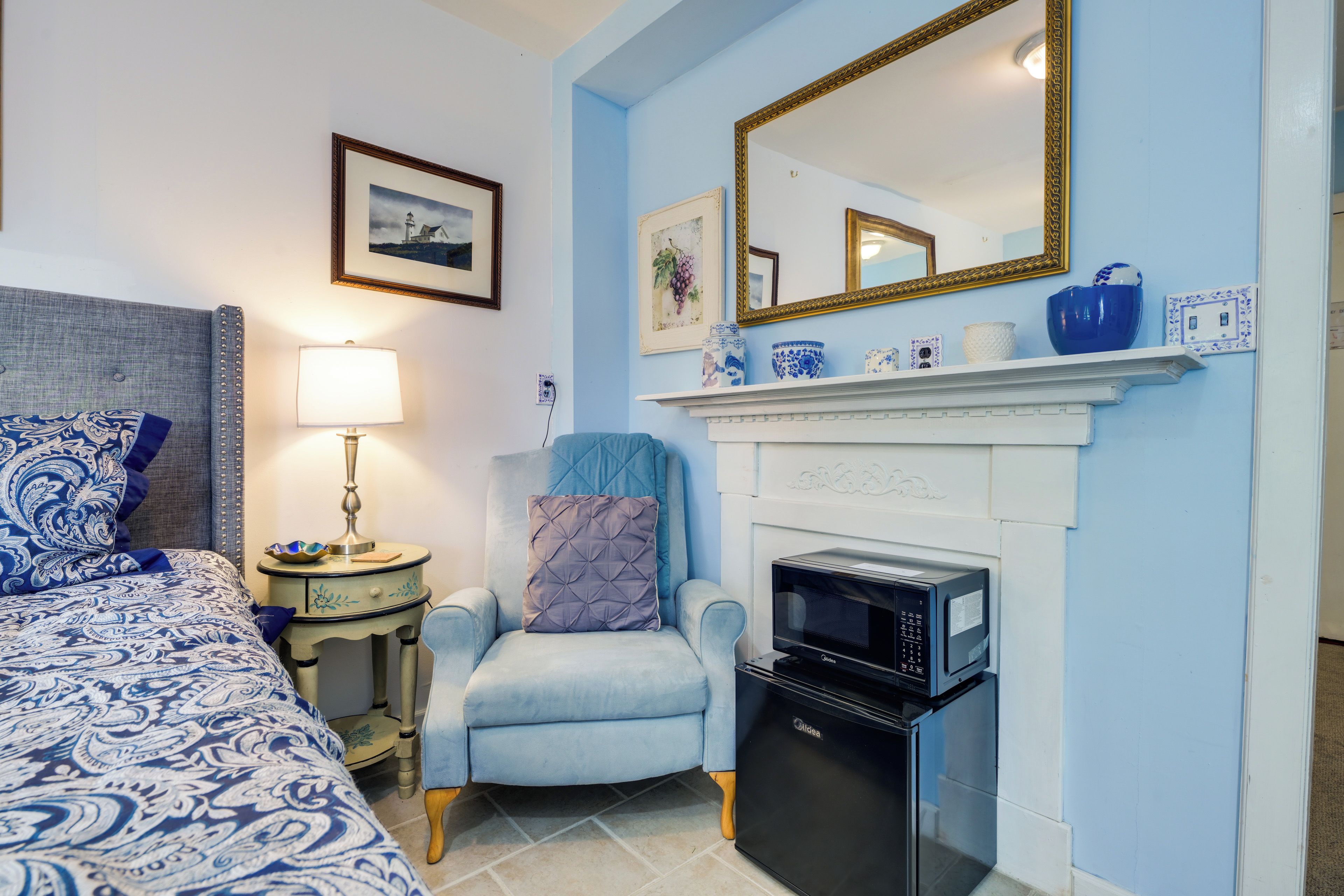 A bedroom with a bed, a chair, a lamp, a fireplace, and a microwave.