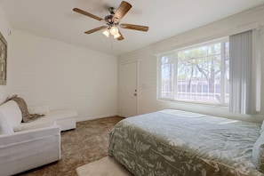 Quarto com cama, sofá branco, ventilador de teto, janela com cortinas e porta.