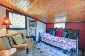 Une chambre avec un plafond en bois, un lit avec une literie à motifs, une chaise, une table et un ventilateur fixé au mur.