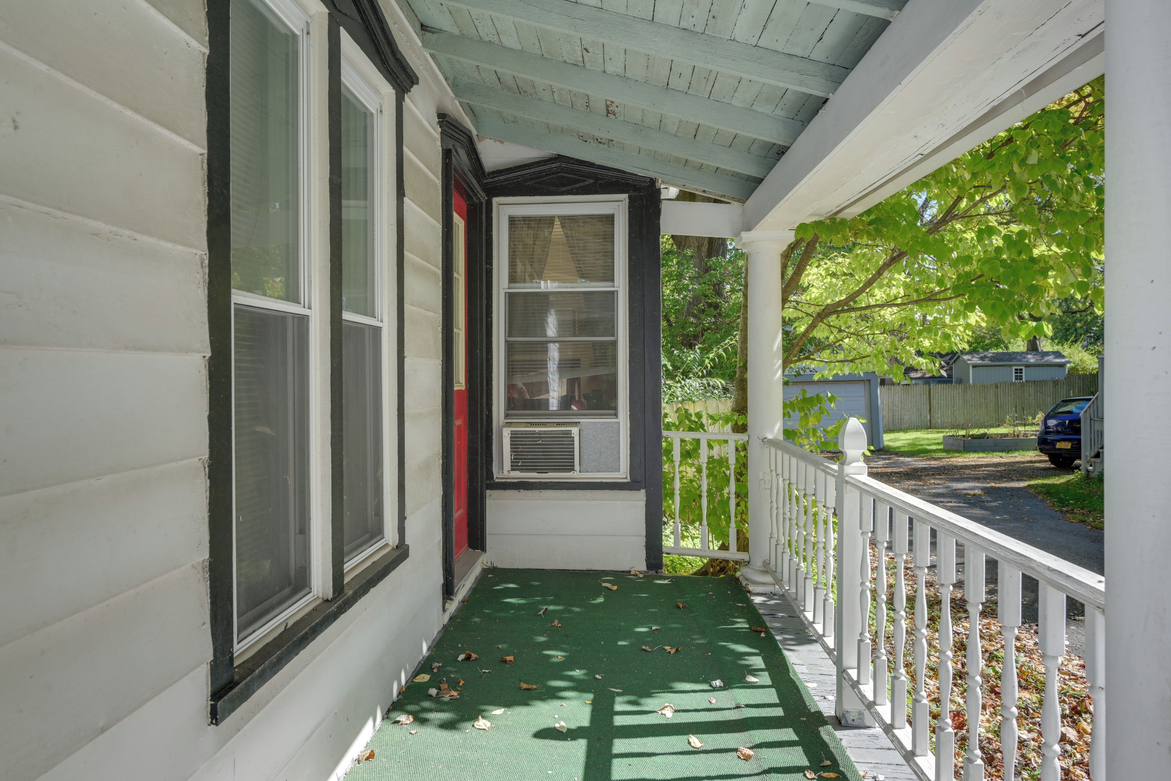 A covered porch with a green mat, white railing, and a view of a residential street.