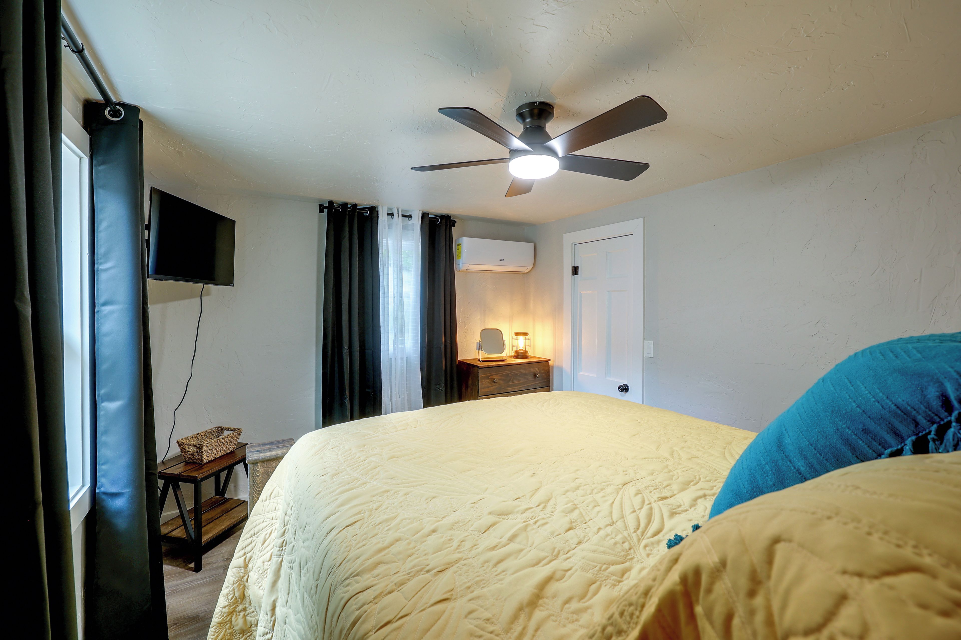 A bedroom with a bed, a ceiling fan, a television, a bedside table with a lamp, and a window with curtains.