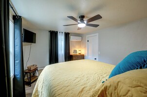 Un dormitorio con cama, ventilador de techo, televisión, mesita de noche con lámpara y ventana con cortinas.