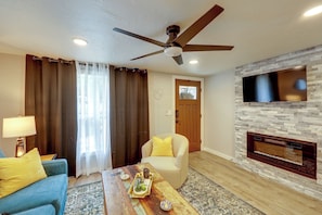 Un soggiorno con ventilatore a soffitto, una TV a schermo piatto montata su una parete in pietra, un divano con cuscini gialli, un tavolino da caffè con oggetti decorativi e una porta con una finestra sopra.