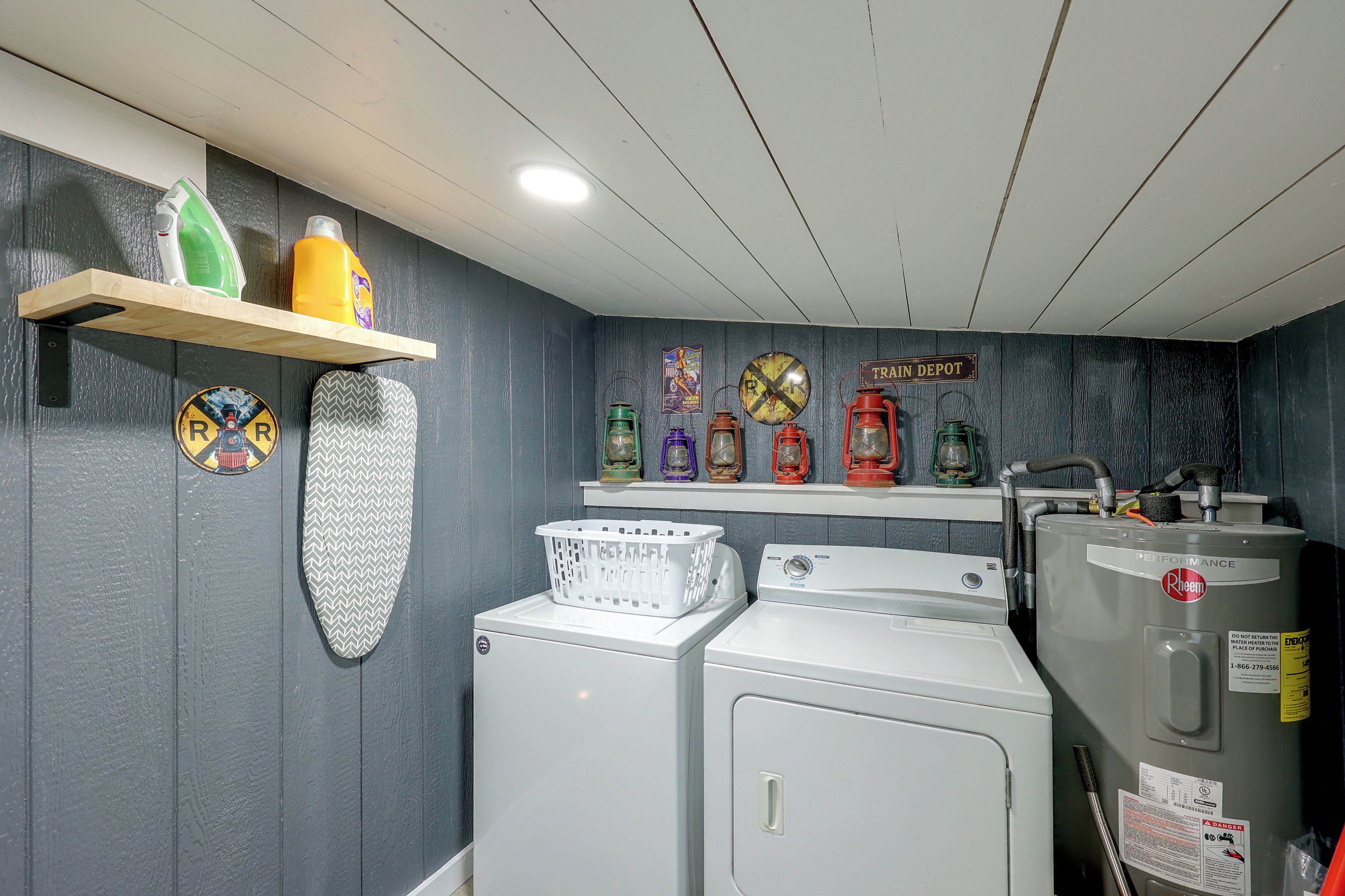 A laundry room with a washing machine, dryer, and ironing board. There are shelves with cleaning supplies and decorative items.