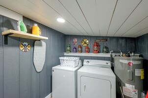 Un cuarto de lavandería con lavadora, secadora y tabla de planchar. Hay estantes con productos de limpieza y objetos decorativos.