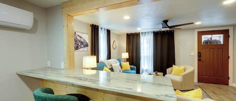A modern kitchen with a marble countertop, wooden bar stools, and a large mirror reflecting the living area.