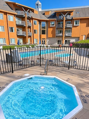 Zona de piscina con jacuzzi, rodeada por una valla, y un edificio de varios pisos con balcones al fondo.