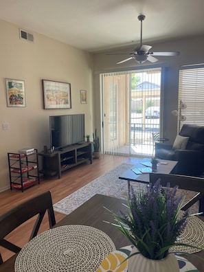 A living room with a television, a ceiling fan, a dining table with chairs, and a sliding glass door leading to a balcony. - Fun in the Sun, Baseball, Luxury Condo! (Surprise)