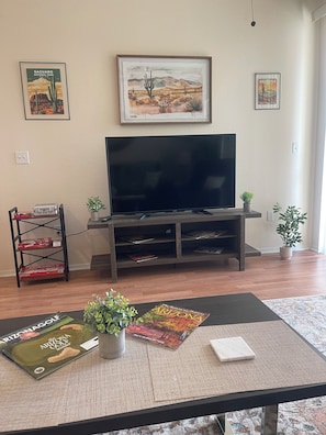 Uma sala de estar com televisão, quadros, uma estante com livros, mesa de centro com revistas e uma planta.