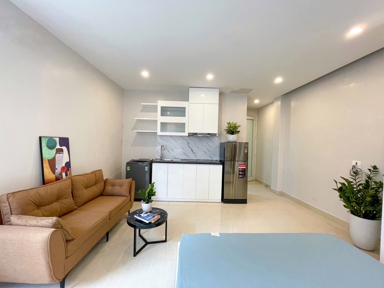 A modern living room with a sofa, a coffee table, and a blue rug. There is a kitchen area with a refrigerator and a sink in the background.