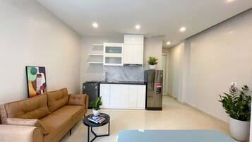 A modern living room with a sofa, a coffee table, and a blue rug. There is a kitchen area with a refrigerator and a sink in the background.