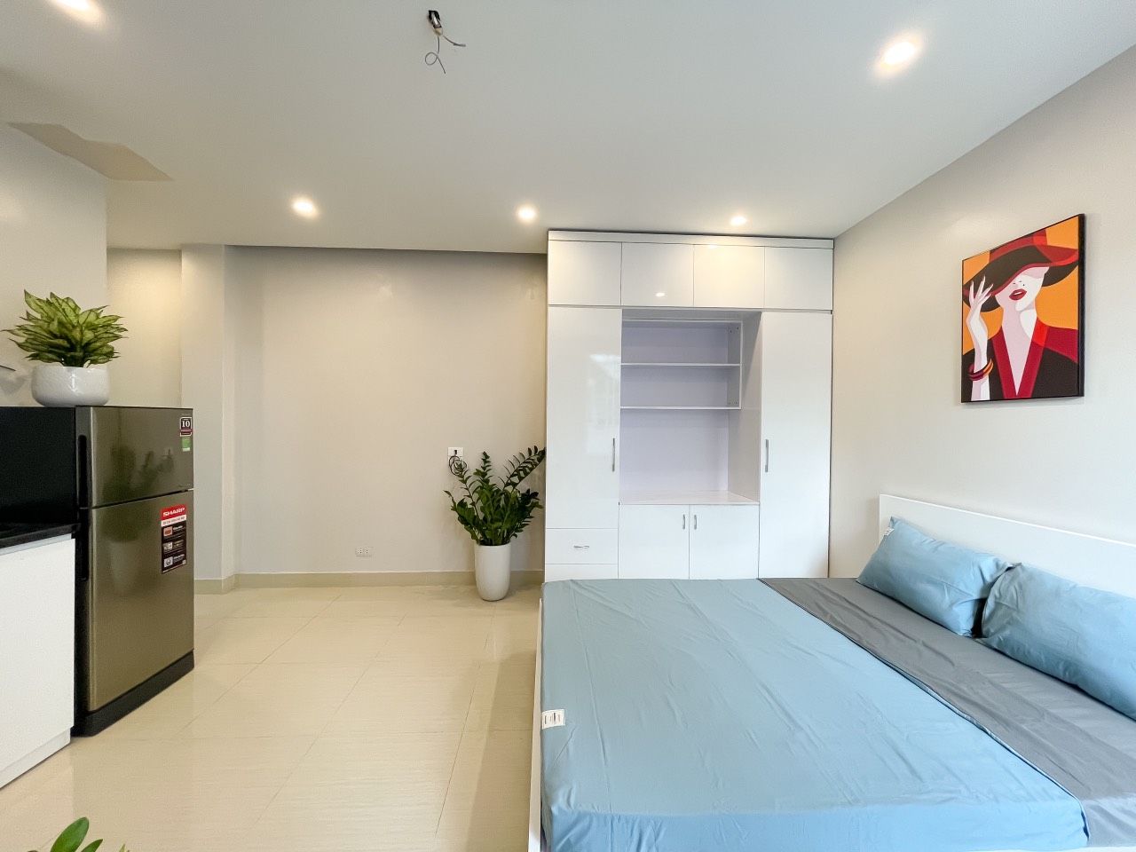 A modern bedroom with a bed, a wardrobe, a small kitchenette, and a potted plant.