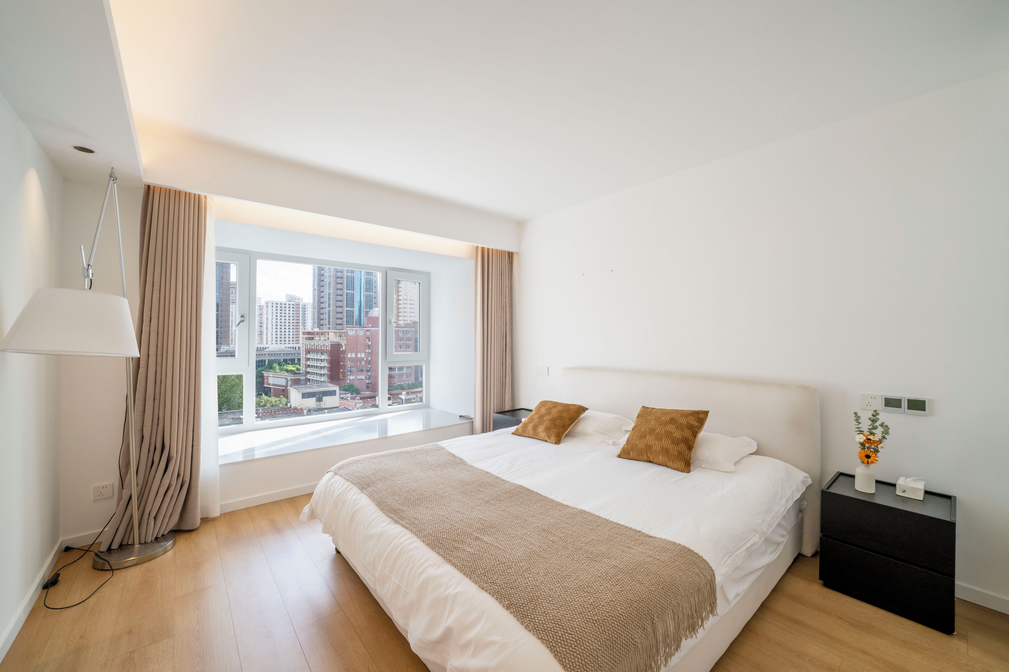 A modern hotel room with a large bed, a bedside table, a lamp, and a view of the cityscape through the window.