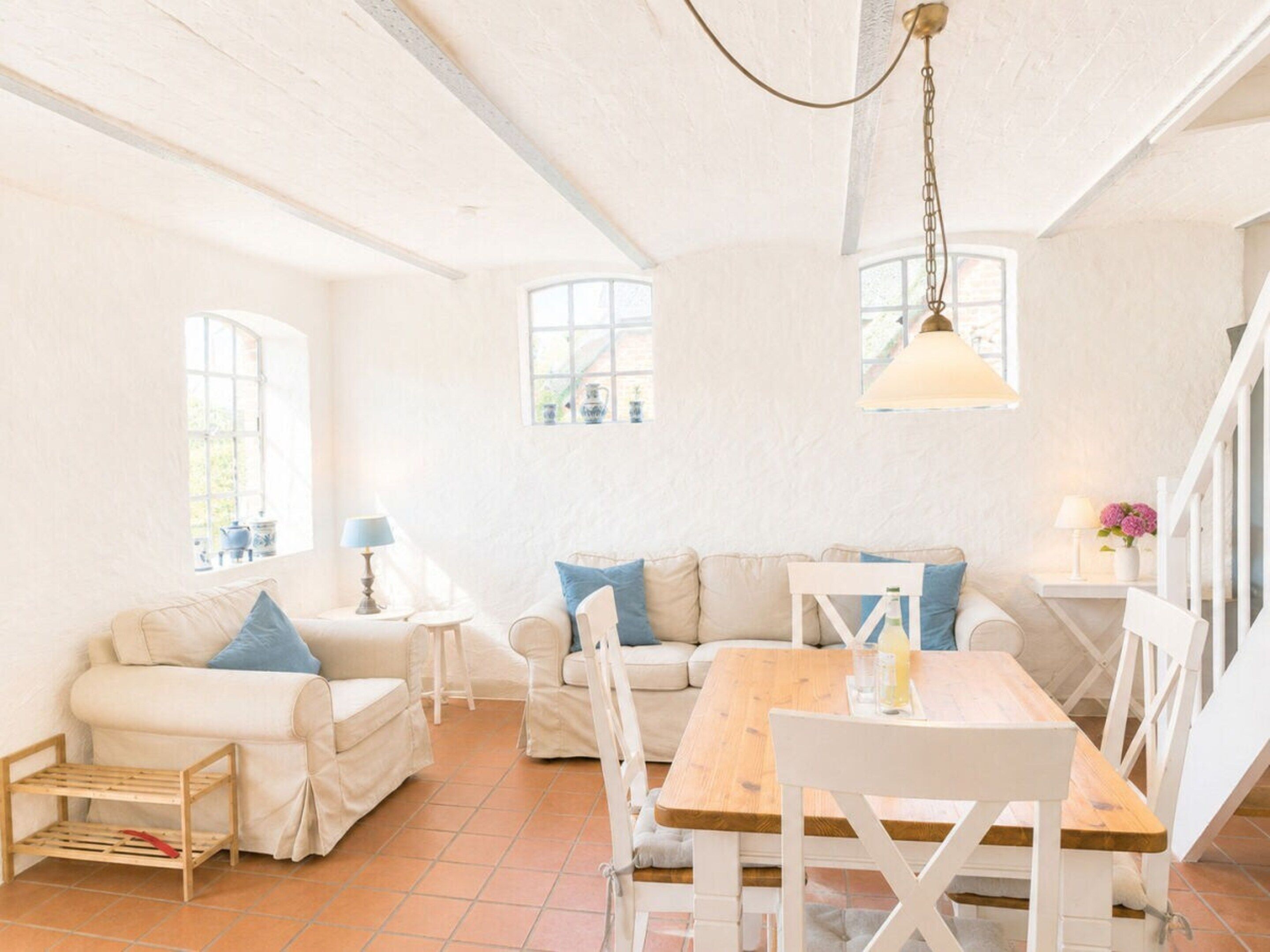 A bright, airy room with a wooden dining table, two white sofas with blue cushions, and a hanging light fixture.