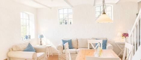 A bright, airy room with a wooden dining table, two white sofas with blue cushions, and a hanging light fixture.