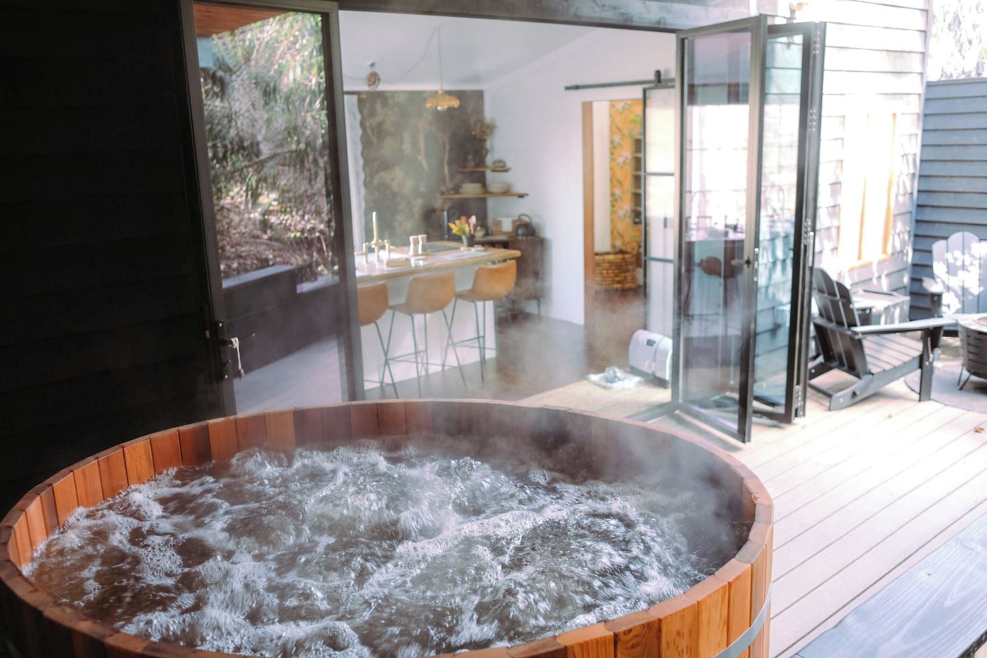 A hot tub with a wooden exterior, a glass door leading to an outdoor seating area with chairs and a table, and a view of a garden through the door.