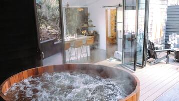 A hot tub with a wooden exterior, a glass door leading to an outdoor seating area with chairs and a table, and a view of a garden through the door.