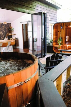 A wooden barrel-shaped hot tub with a metal railing in front, a dining area with chairs and a table, and a glass door leading to an outdoor space. - Dark City Solarium – Sauna + Hot Tub + Luxury (Black Mountain)