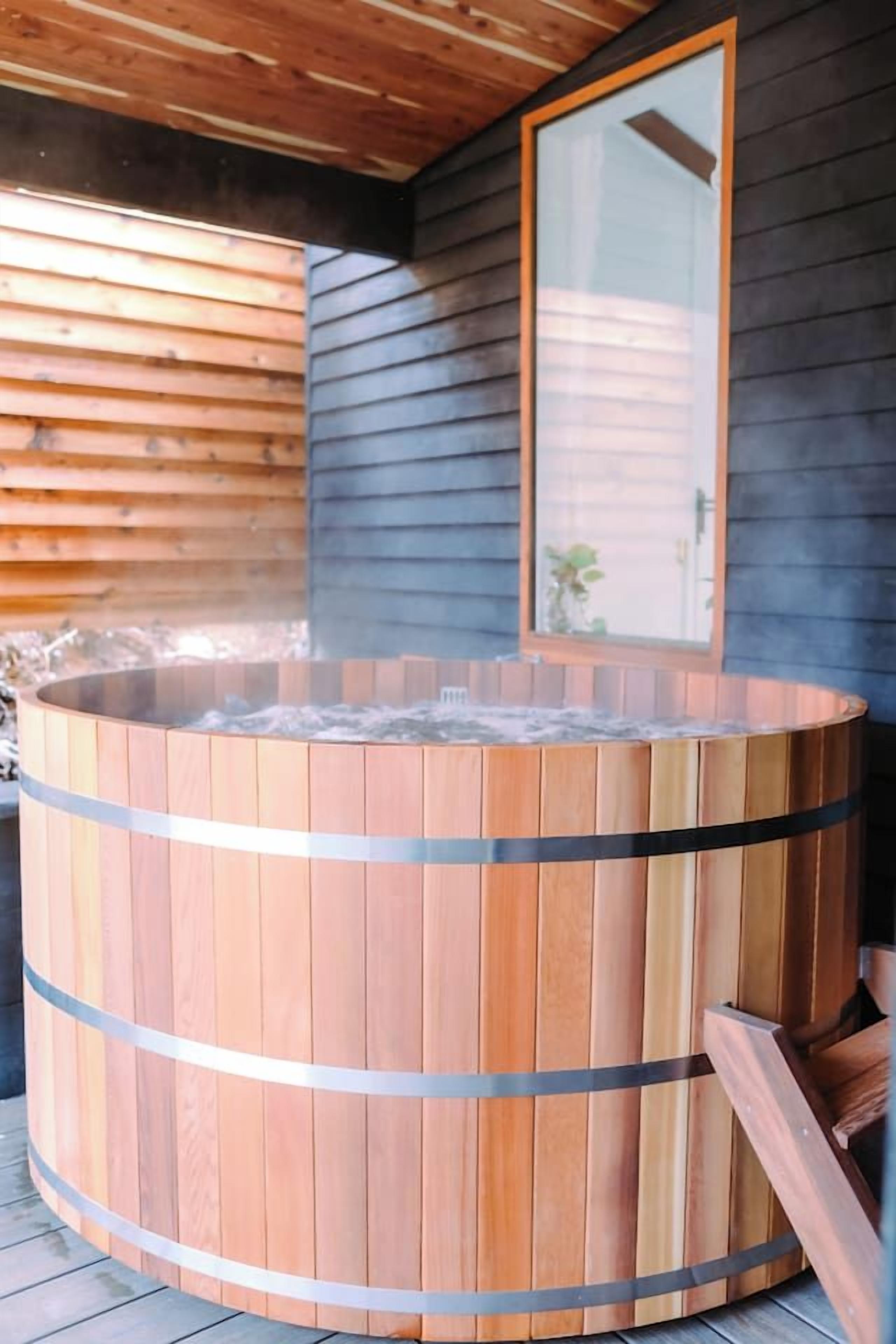 Una bañera de madera con bandas metálicas, una terraza de madera y una pared exterior oscura con un ventanal.