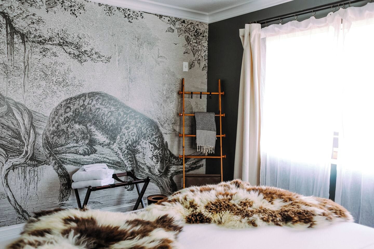 A bedroom with a large wall mural, a bed with a fur throw, a wooden ladder, and a chair.