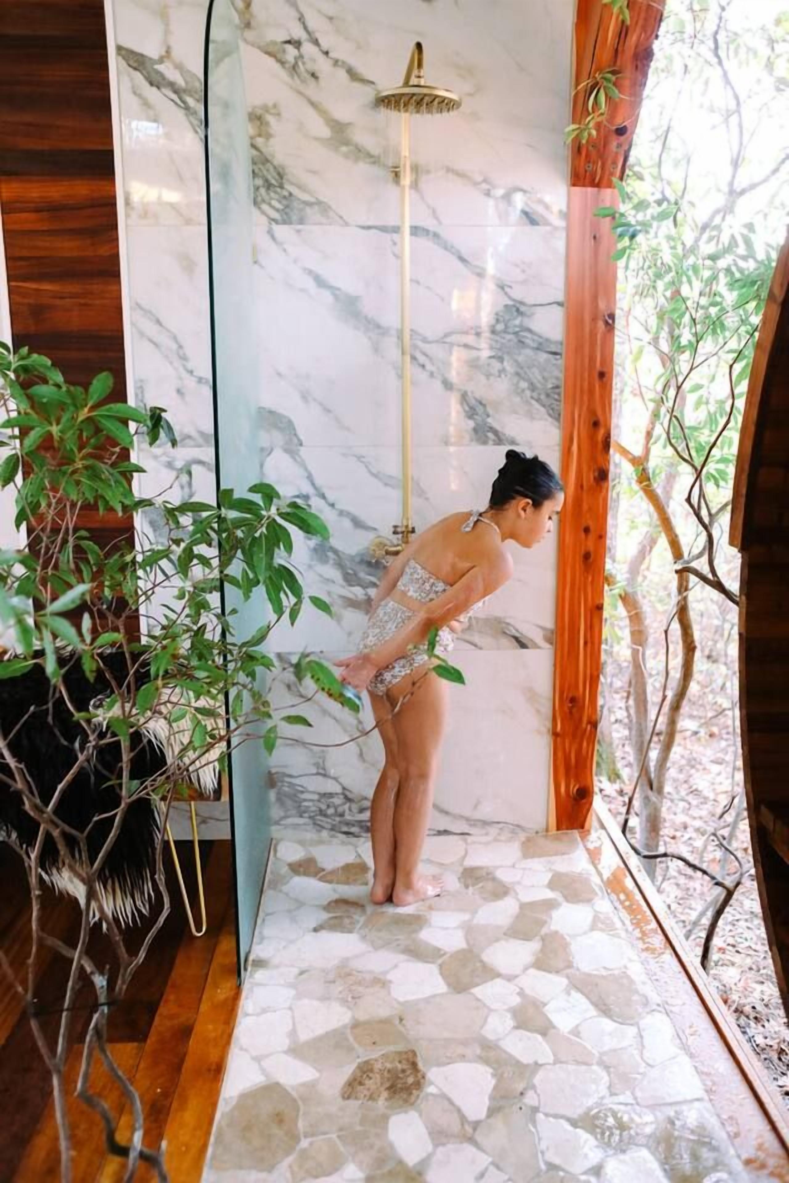 A person in a bathroom with a marble wall, a shower, and a wooden floor.