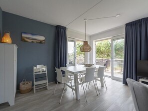 A modern kitchen with white cabinetry, a dining table with chairs, a hanging light fixture, and a large window with curtains. - Modern 5-guest Holiday Apartment With Balcony (Julianadorp)