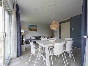 A modern dining area with a white table and chairs, a hanging light fixture, and a view of the outdoors through a large window. - Modern 5-guest Holiday Apartment With Balcony (Julianadorp)