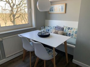 A room with a white table, two white chairs, a radiator, a window with a view of trees, and a bench with patterned cushions. - Jaardenhuug - OG (Utersum)