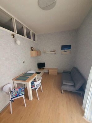 Un ambiente compatto con pavimento in legno, un divano grigio, un tavolino con sedie, una televisione e uno scaffale con oggetti decorativi.