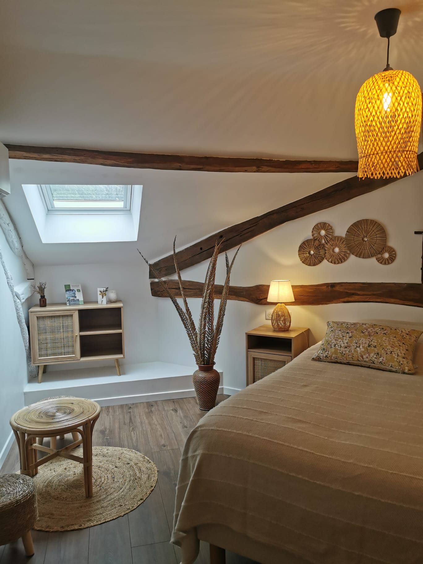 Un dormitorio acogedor con techo inclinado, tragaluz, cama, mesita de noche con lámpara, pequeño taburete y cesta tejida.