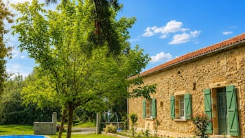 Un edificio de piedra con contraventanas verdes, una piscina y mobiliario exterior.