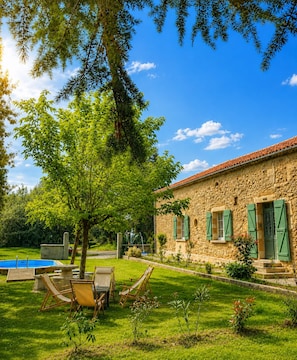Un edificio de piedra con contraventanas verdes, una piscina y mobiliario exterior.