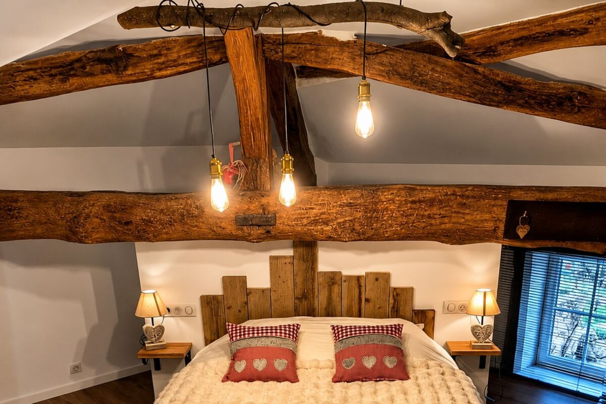Un dormitorio con techo de madera, una cama con manta beige suave, dos almohadas con estampado de corazones, lámparas de noche y una ventana con persianas.