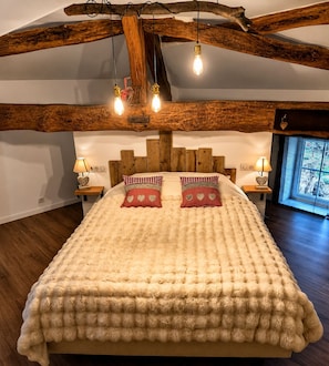 Un dormitorio con techo de madera, una cama con manta beige suave, dos almohadas con estampado de corazones, lámparas de noche y una ventana con persianas.