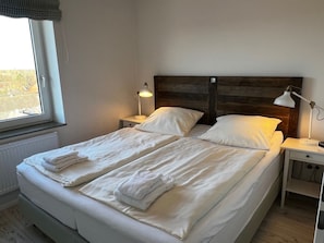 A double bed with white linens and towels, a wooden headboard, bedside tables with lamps, and a window with a view of buildings. - Holiday Apartment in Wyk on Fohr With Balcony (Wyk auf Foehr)