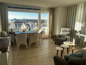 A modern living room with a dining area, large windows, and a view of a marina. - Holiday Apartment in Wyk on Fohr With Balcony (Wyk auf Foehr)