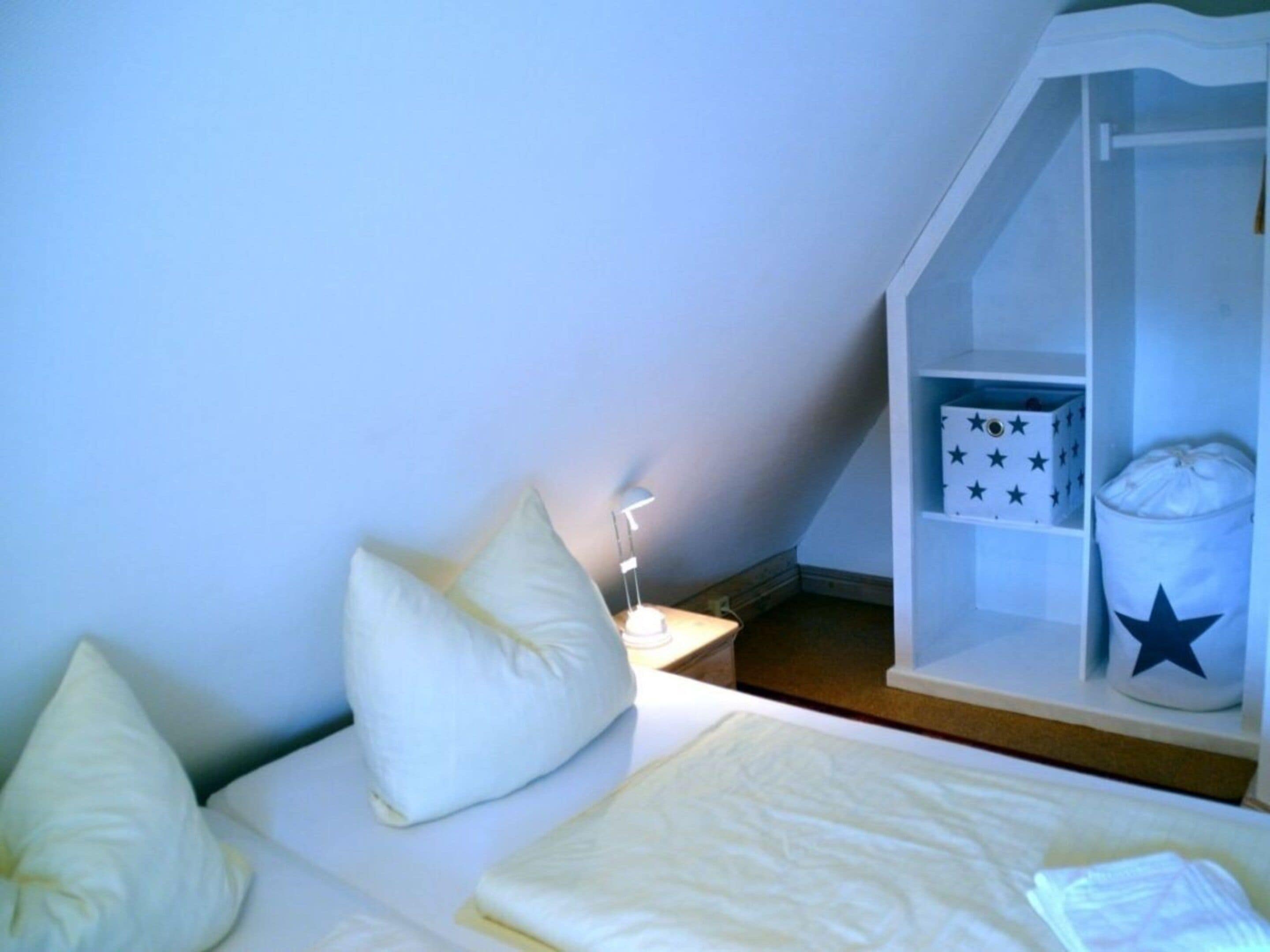 Une chambre à coucher comprenant un lit, des oreillers, une table de chevet, une lampe, une étagère avec des boîtes de rangement et une poubelle.
