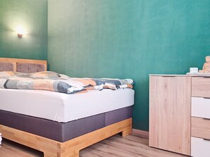 Uma cama com estrutura de madeira e colchão escuro, uma cômoda de madeira com gavetas e uma parede verde com luminária.