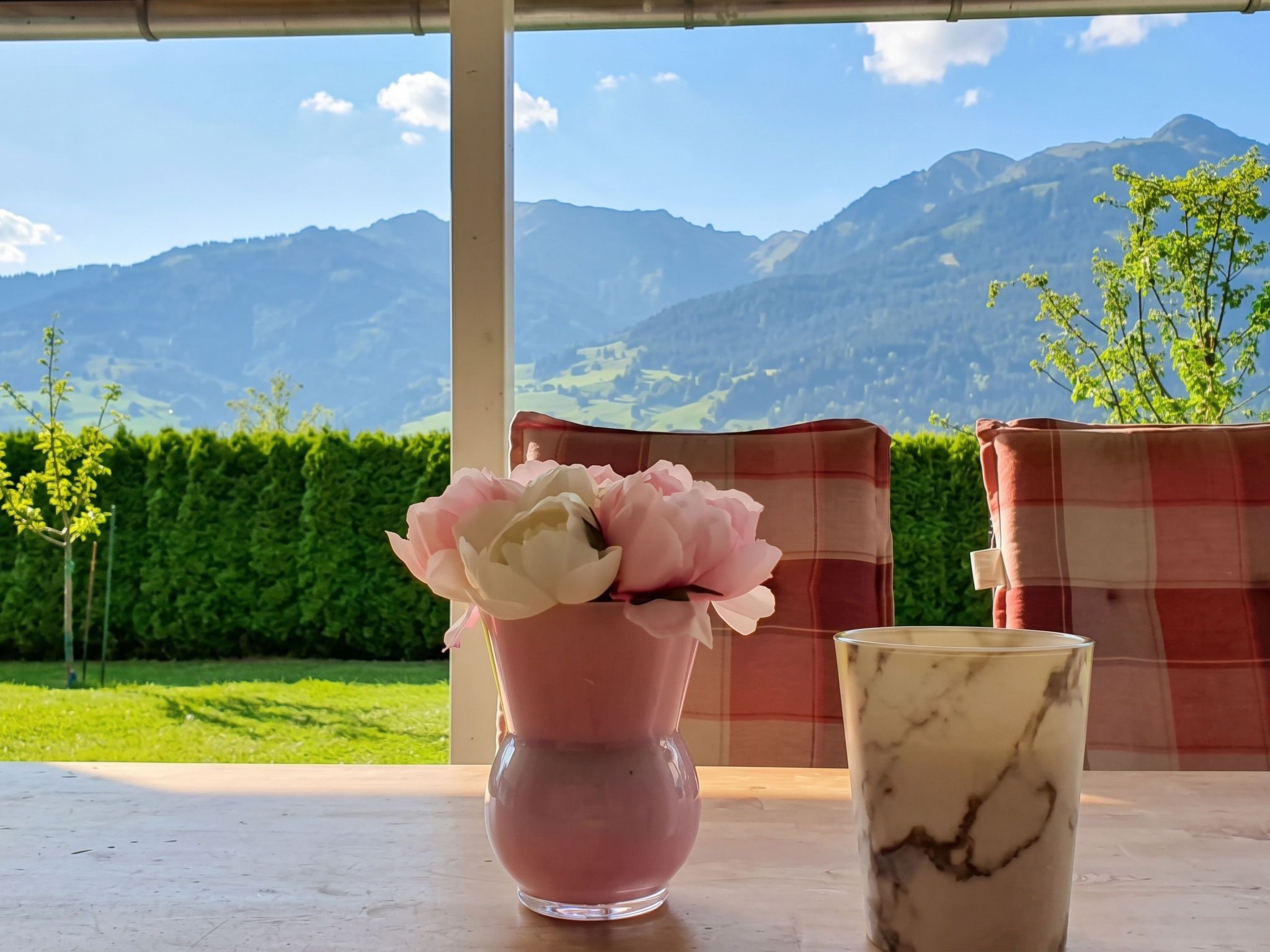 A table with a vase of flowers and a glass, set against a backdrop of mountains and a clear sky.