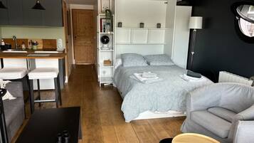 Un espace de vie compact comprenant un coin cuisine, un lit avec une couette à motifs, un canapé gris et une petite table ronde en bois.