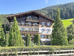 Et hotel med flere etager, med altaner, omgivet af grønt og med et bjergrigt landskab i baggrunden.