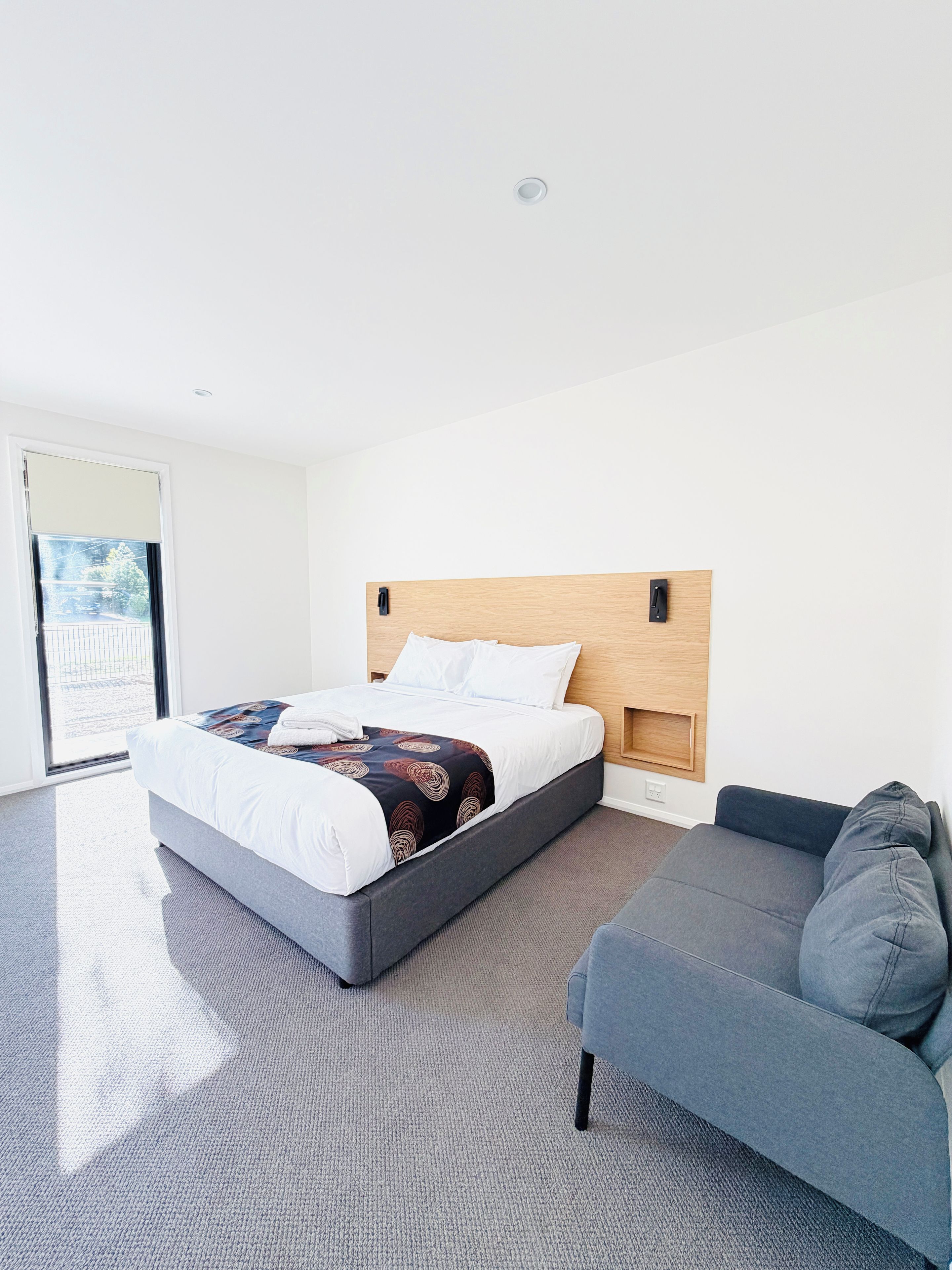 A modern hotel room with a bed, a grey sofa, and a large window.