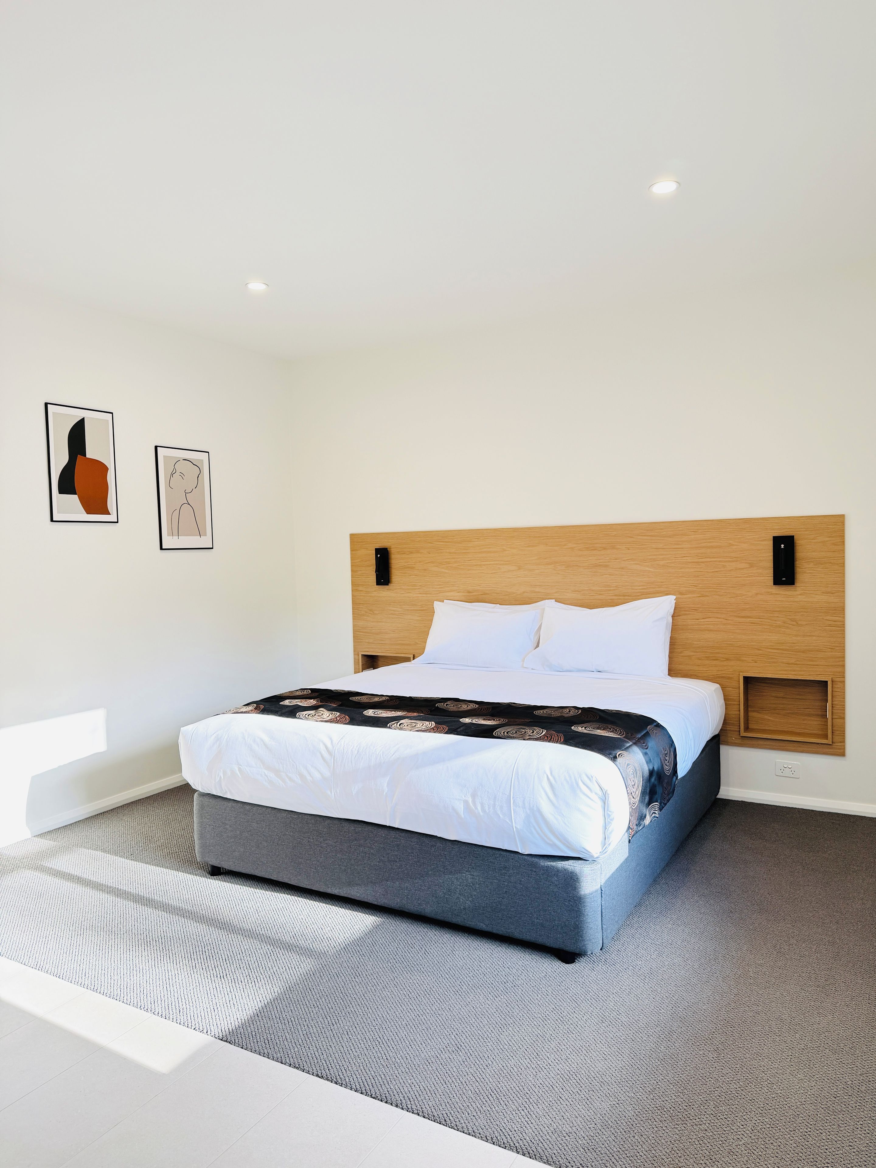 A modern hotel room with a large bed, a wooden headboard, and two framed artworks on the wall.