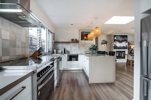 A modern kitchen with a central island, stainless steel appliances, and a dining area with a view. - Cottesloe Coastal Escape | Fire Pit & Games (Barwon Heads)