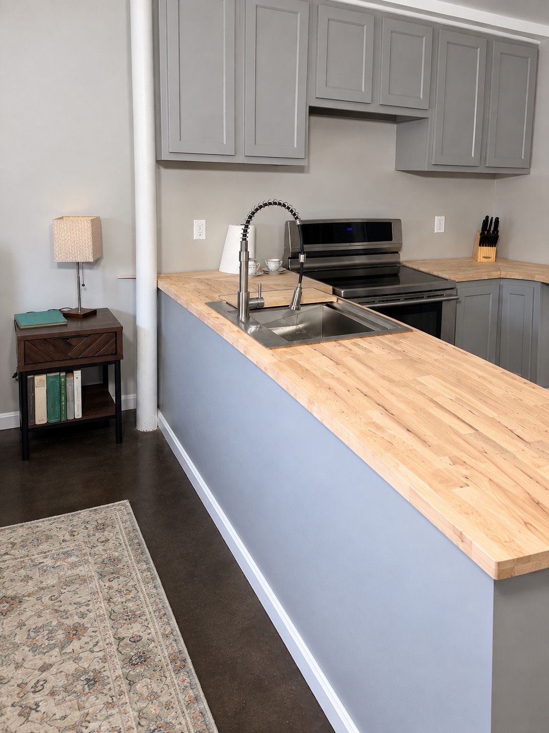 Una cocina con armarios grises, encimera de madera, pileta de acero inoxidable, estufa y una pequeña mesa con lámpara y libros.